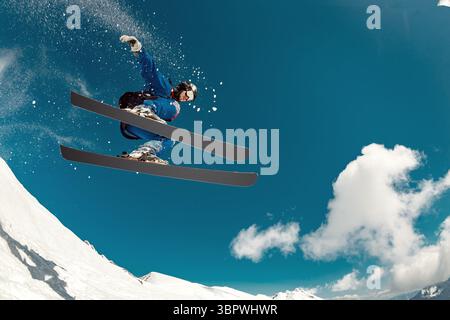 A shot from below of a skier performing a high extreme jump while off-piste skiing against a snowy slope and clear blue sky. Dynamic flight, adrenaline and freedom of freeride Stock Photo