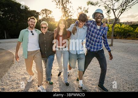 Full length shot of a happy and diverse group of young friends with their arms around each other, walking towards the camera and laughing together in Stock Photo