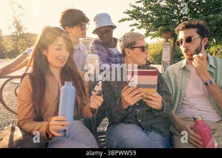 A creative team of diverse young students working together on a laptop and tablet while sitting on a park bench, having a brainstorming session for a Stock Photo