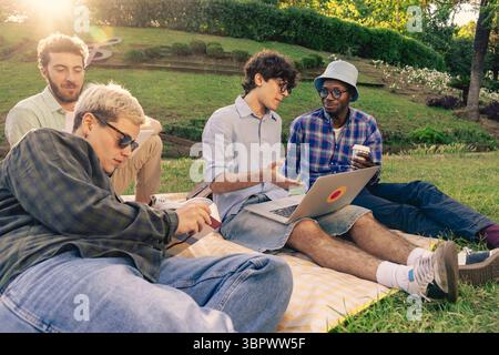 A multiethnic group of friends or students relaxing on a blanket in a park, studying and working together with a laptop on a sunny day with beautiful Stock Photo