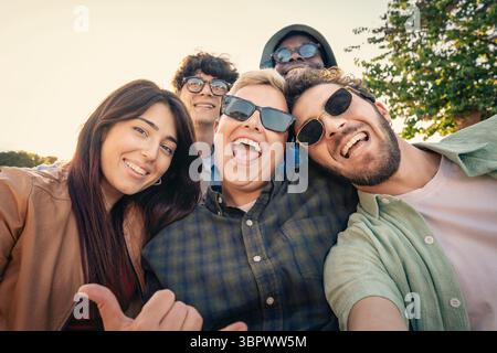 A cheerful and diverse multiethnic group of young adult friends laughing and smiling at the camera, taking a fun selfie together outdoors during a bea Stock Photo