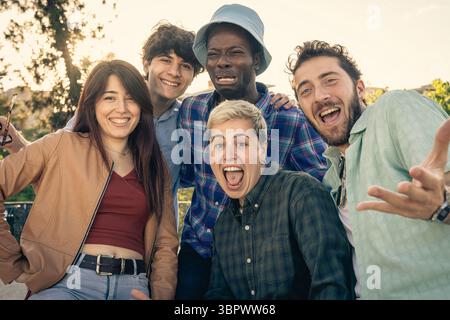 An energetic and diverse multiethnic group of friends makes funny and exaggerated faces while joking and laughing together outdoors, showing a moment Stock Photo