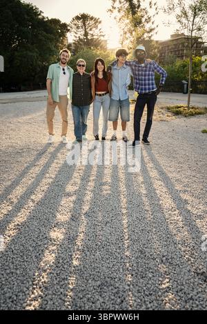 A diverse multiethnic group of friends or colleagues stands together with their arms around each other, posing for a team portrait outdoors at sunset Stock Photo