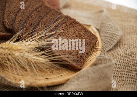 Slices of fresh dark rye bread in a white bowl with wheat ears on rustic burlap background. Healthy traditional bakery product. Stock Photo
