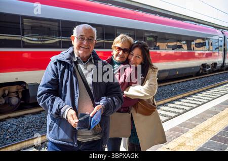 Smiling elderly couple with their adult daughter standing on a train platform. The elderly man holds a smartphone and wallet, while the women stand be Stock Photo