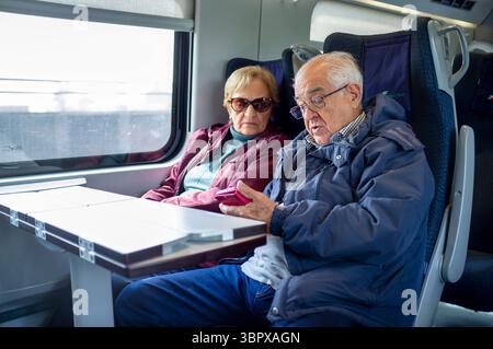 Elderly couple sitting side by side on a modern train, looking at a smartphone together. The man is showing something on the screen while the woman li Stock Photo