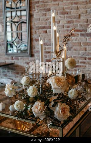 A refined floral arrangement with roses and candles on a mirrored table, set against a brick wall. the warm lighting creates a cozy, elegant ambiance. Stock Photo