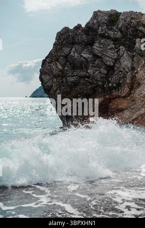 Waves crashing against a large rocky cliff by the sea under a clear sky. Stock Photo