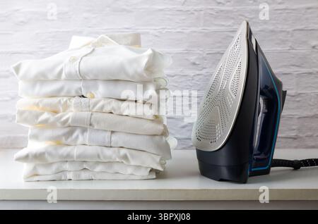 stack of white shirts on a white wooden cabinet with an iron next to it in an ironing room. Front view. Stock Photo