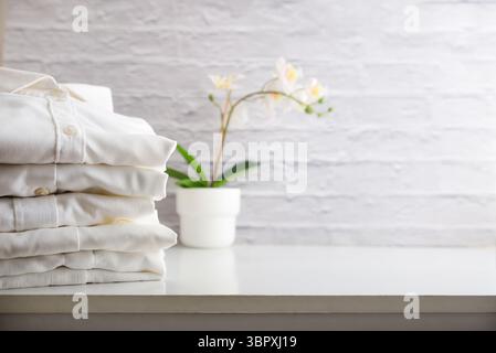 stack of clean, ironed, folded, and freshly laundered white shirts on white wooden furniture with orchid in the background and white brick wall. Front Stock Photo