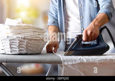 Detail of an elegant man ironing a white shirt on an ironing board with a basket filled with a stack of freshly ironed shirts in a living room. Front Stock Photo
