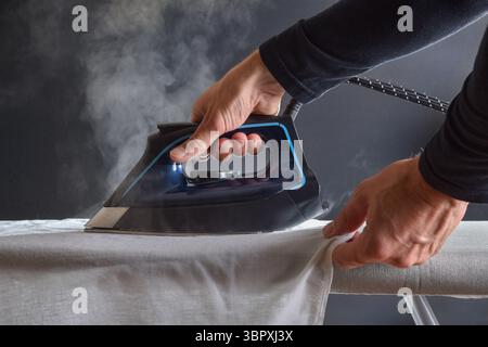 Detail of man ironing a white shirt with a steam iron and dark isolated background. Front view. Stock Photo