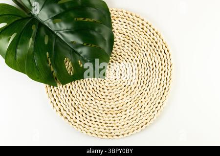 Top view of brown wicker table mat, green monstera leaf on white background. Straw round tray, flat lay, copy space. Stock Photo