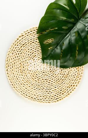 Top view of brown wicker table mat, green monstera leaf on white background. Straw round tray, flat lay, copy space. Stock Photo