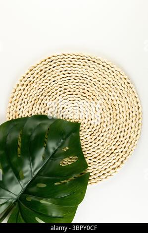 Top view of brown wicker table mat, green monstera leaf on white background. Straw round tray, flat lay, copy space. Stock Photo