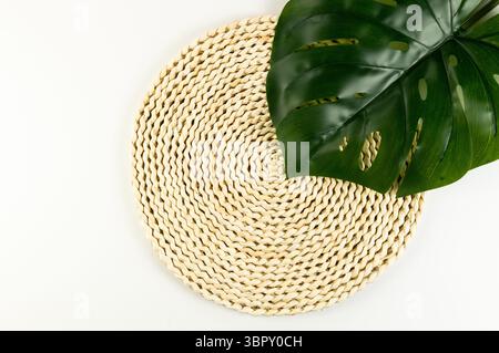 Top view of brown wicker table mat, green monstera leaf on white background. Straw round tray, flat lay, copy space. Stock Photo