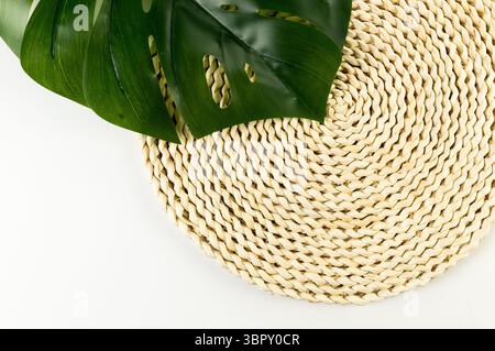 Top view of brown wicker table mat, green monstera leaf on white background. Straw round tray, flat lay, copy space. Stock Photo