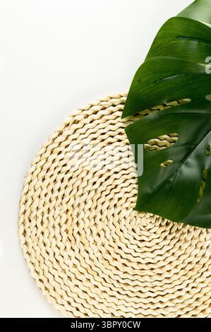 Top view of brown wicker table mat, green monstera leaf on white background. Straw round tray, flat lay, copy space. Stock Photo