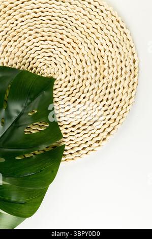 Top view of brown wicker table mat, green monstera leaf on white background. Straw round tray, flat lay, copy space. Stock Photo