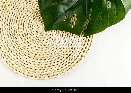 Top view of brown wicker table mat, green monstera leaf on white background. Straw round tray, flat lay, copy space. Stock Photo