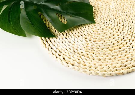 Top view of brown wicker table mat, green monstera leaf on white background. Straw round tray, flat lay, copy space. Stock Photo
