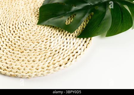 Top view of brown wicker table mat, green monstera leaf on white background. Straw round tray, flat lay, copy space. Stock Photo