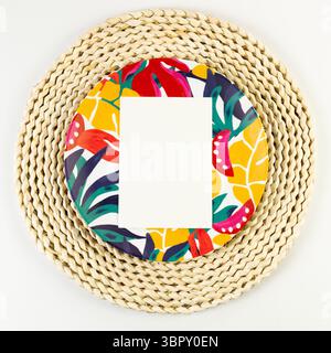Top view of brown wicker table mat, white card, colorful plate on white background. Straw round tray, exotic plant plate, summer picnic. Stock Photo