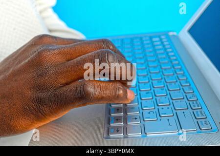 Close-up of dark-skinned programmer typing on laptop keyboard, focused on software development against a vibrant blue backdrop Stock Photo