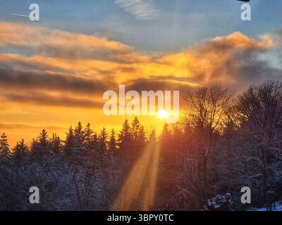 Beautiful sun rays and a lens flare burst from behind a silhouetted forest hilltop, creating an inspirational and hopeful scene during a winter sunset Stock Photo