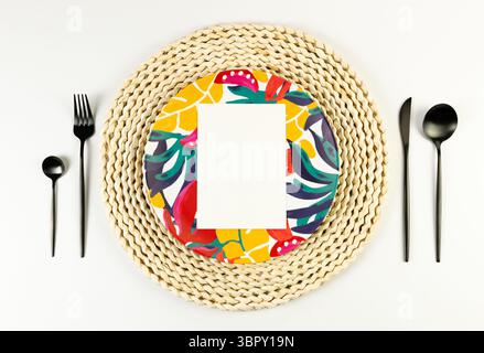 Top view of brown wicker table mat, colorful plate, black cutlery on white background. White card, straw round tray, exotic plant plate, summer picnic. Stock Photo