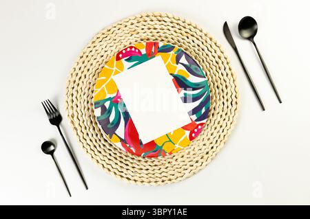 Top view of brown wicker table mat, colorful plate, black cutlery on white background. White card, straw round tray, exotic plant plate, summer picnic. Stock Photo