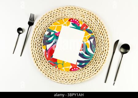 Top view of brown wicker table mat, colorful plate, black cutlery on white background. White card, straw round tray, exotic plant plate, summer picnic. Stock Photo