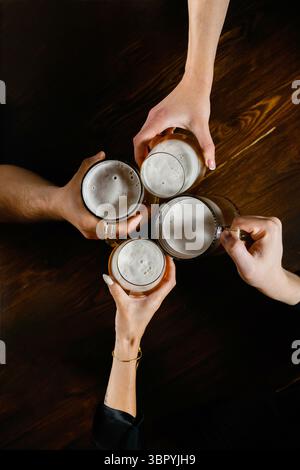 Top view of four hands clinking beer mugs over a dark wooden table. Foamy light beer in clear glass mugs. Two men and two women participating in a cel Stock Photo