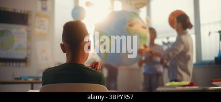 STEM Class on Renewable Energy: Female Teacher Explaining Energy Resources to Elementary School Students Using Earth Model. Rack Focus on African American Boy Learning Science in Classroom. Dolly Shot Stock Photo