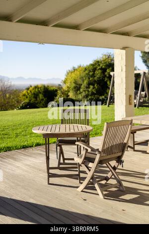 Round wooden table and two folding chairs resting on deck under white patio roof, mountain backdrop. Rustic, outdoor, leisure, natural, serene, relaxi Stock Photo
