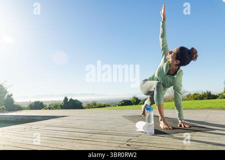 Mature woman in sportswear twisting yoga on pool deck with yoga mat, water bottle, copy space. Wellness, outdoor, posture, serenity, relaxation, natur Stock Photo