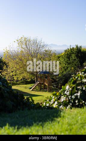 Wooden playset featuring slide and ladder is standing on sloping grassy backyard with mountain view. Playground, outdoor, rustic, leisure, nature, adv Stock Photo