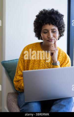 African American woman in yellow sweater sitting on teal cushion by window typing on silver laptop. Casual, workspace, modern, comfort, concentration, Stock Photo