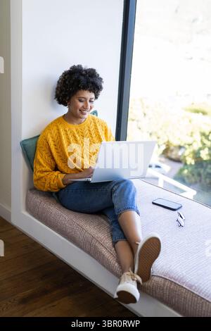 African American woman sitting on window seat in reading nook, using laptop with smartphone. Cozy, leisure, workspace, digital, nature, lifestyle, mod Stock Photo