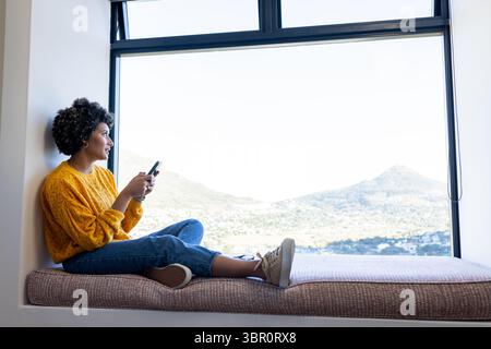 African American female in sweater sitting on window seat holding phone gazing at view, copy space. Contemplation, modern, cozy, scenic, tranquility, Stock Photo
