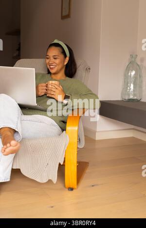 Asian woman sitting on armchair in living room wearing green sweater holding mug and viewing laptop. Cozy, relaxation, homeoffice, minimalist, lifesty Stock Photo