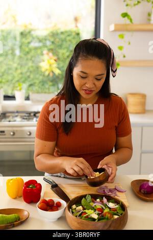 Asian woman slicing onion with chef's knife on wooden board while preparing salad at kitchen island. Healthy eating, culinary, organic, lifestyle, min Stock Photo