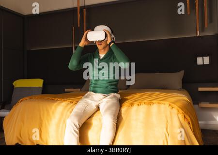 Man is wearing VR headset and is sitting on bed with mustard blanket, copper pendant lights. Technology, relaxation, modern, ambient, leisure, interio Stock Photo