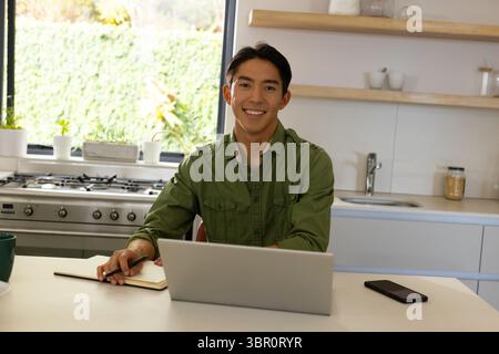Asian man sitting at kitchen island working on laptop with notebook pen smartphone and coffee mug. Modern, workspace, minimalist, wellness, productivi Stock Photo