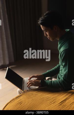 Asian man wearing green sweater sitting on mustard sofa typing on laptop at home with smartwatch. Cozy, modern, corporate, workspace, interior, focuse Stock Photo