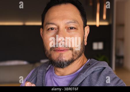 Asian man wearing gray hoodie sitting in modern living room with beige sofa and pendant lights. Portrait, masculinity, contemporary, relaxation, home Stock Photo