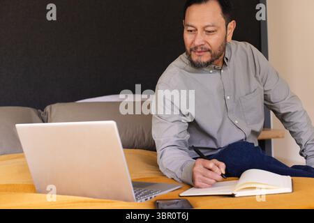 Asian man writing in notebook beside laptop while sitting on bed in bedroom workspace, copy space. Concentration, productivity, homeoffice, workspace, Stock Photo