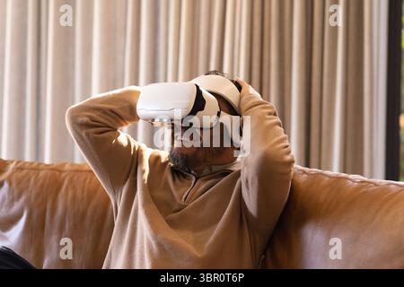 Asian man adjusting white VR headset on brown leather sofa by beige curtains in living room. Technology, relaxation, modern, comfort, leisure, lifesty Stock Photo