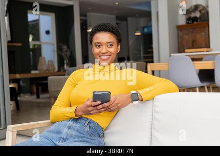 African American woman sitting on white sofa at home wearing smartwatch holding smartphone smiling. Modern, relaxation, cozy, casual, lifestyle, home, Stock Photo