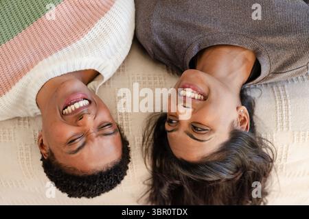 Diverse couple lying side by side on cream-colored woven blanket at home and smiling together. Romance, relaxation, intimacy, cozy, connection, love, Stock Photo